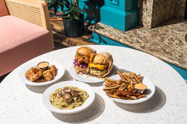 The image shows a round table with burgers, fries, pasta with mushrooms, and fried snacks, all served with dips.
