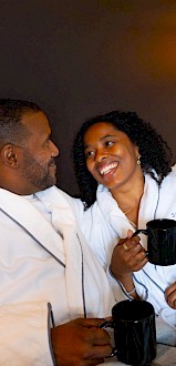 Two people in bathrobes cozy in bed, sharing a smile over mugs of tea, cuddled under a blanket in a warm, intimate hotel scene.
