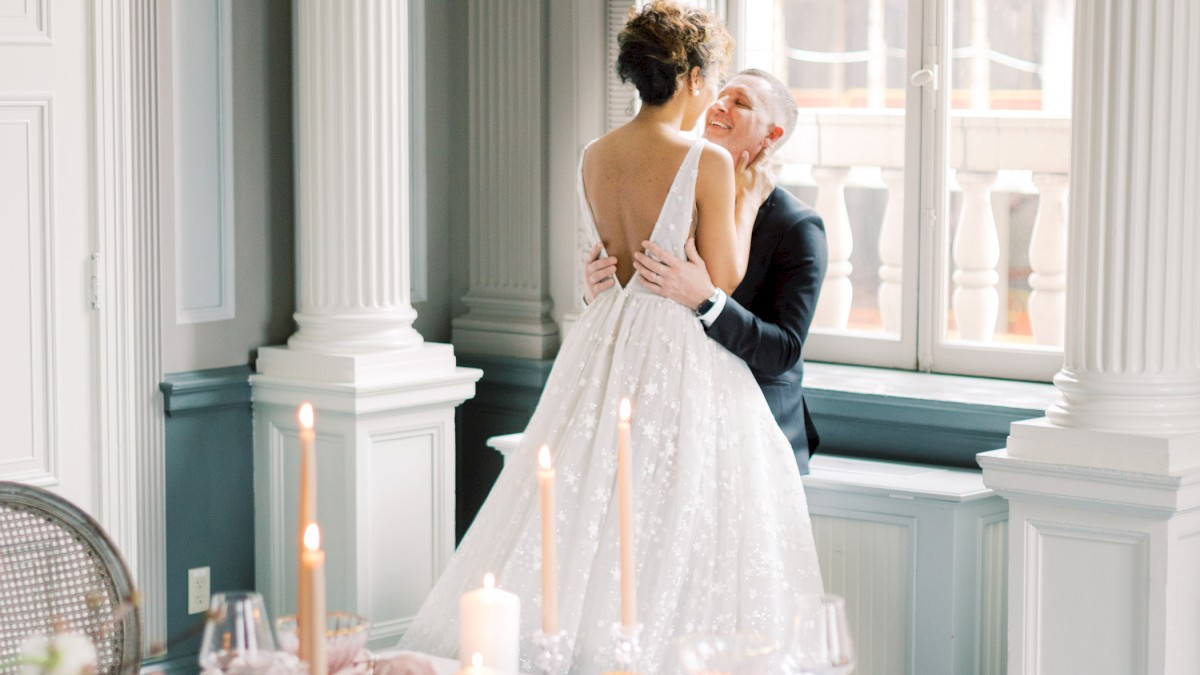 Two people dancing closely in a sunlit, elegantly set dining room with a white-lace wedding dress on the guest. (140 chars, ends with a period.)