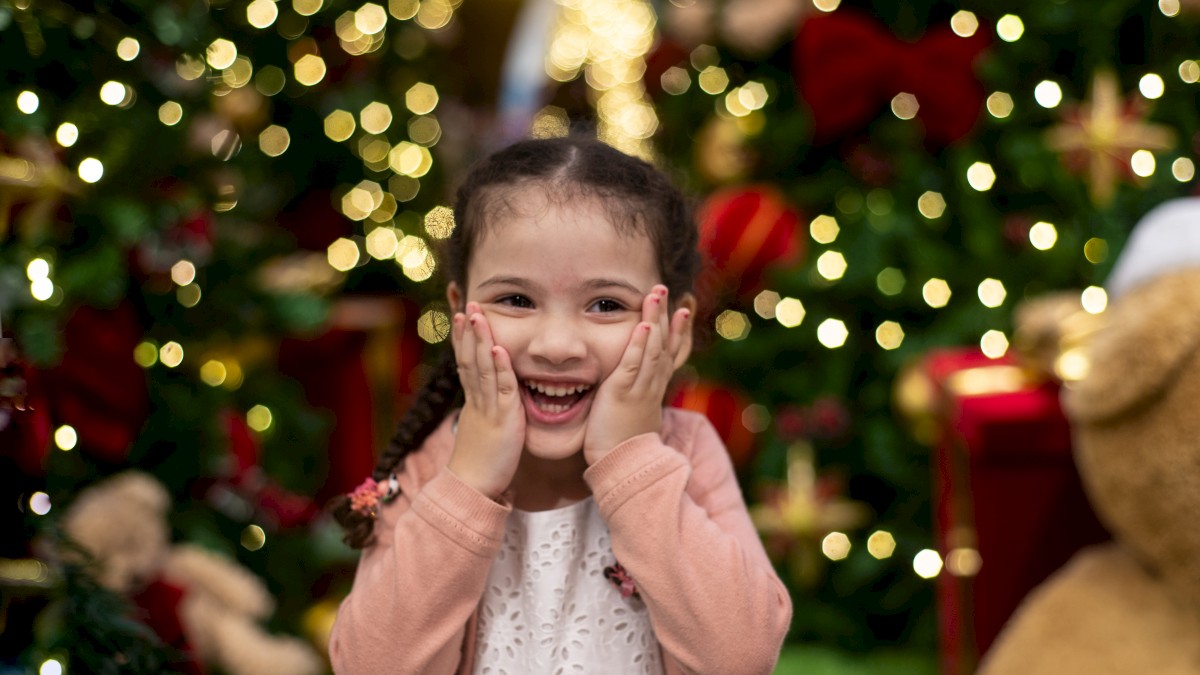 A delighted young girl with braided hair stands in front of a Christmas tree, hands on her cheeks, smiling brightly.