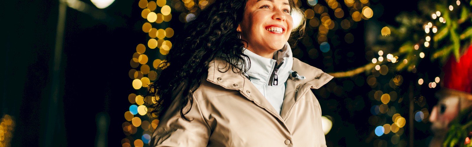 A smiling woman in a beige coat walks joyfully through a festive night market with twinkling lights overhead, enjoying the holiday mood.