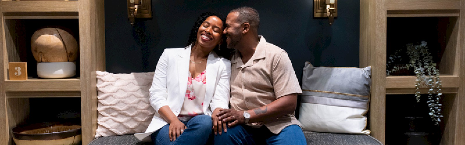 A couple sits closely on a built-in bench in a cozy, shelved room with lamps, cushions, and decorative items around them. End.