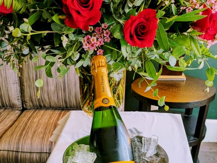 A bottle of champagne chilling in an ice bucket on a silver tray, flutes ready, chocolates in blue boxes, with a lush red flower bouquet backdrop.