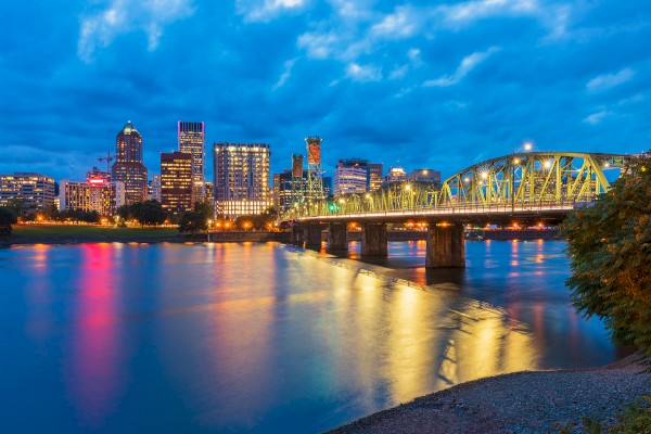 A city skyline at dusk with illuminated buildings and a lit bridge reflecting on a calm river.