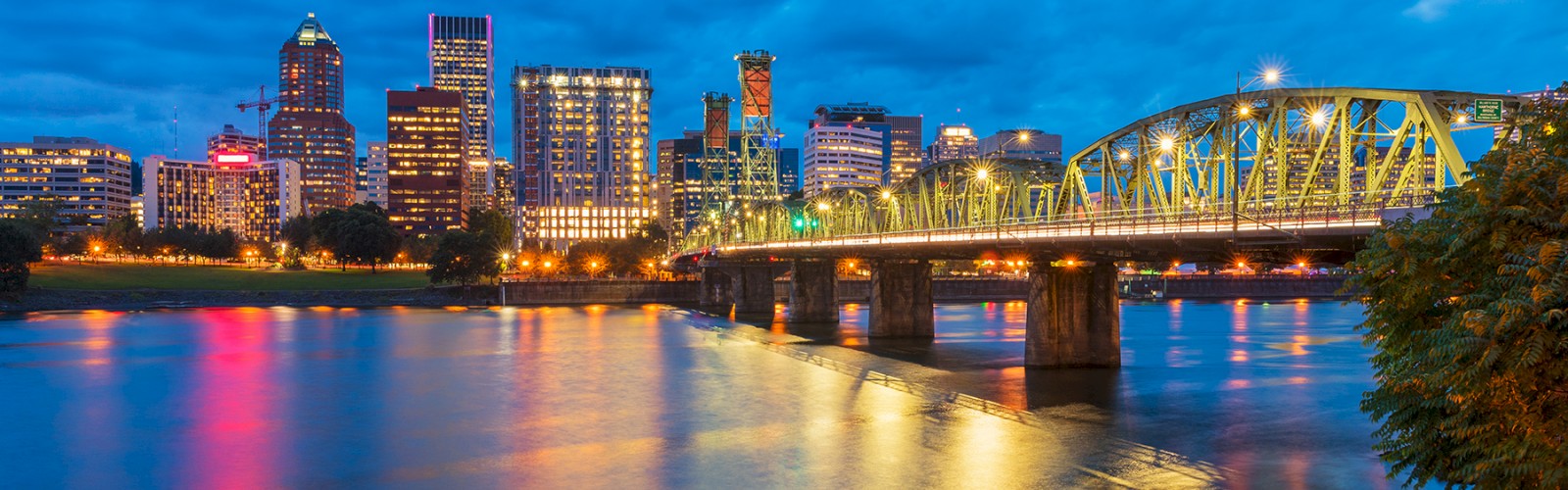 A city skyline at dusk with illuminated buildings, a bridge over a river, and a partly cloudy sky in vibrant colors.