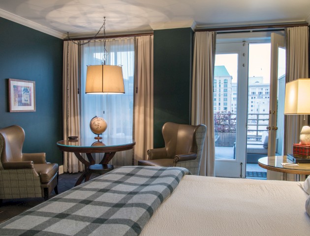 A cozy hotel room with a bed, two wingback chairs, a small table with a globe, and city view through open balcony doors.