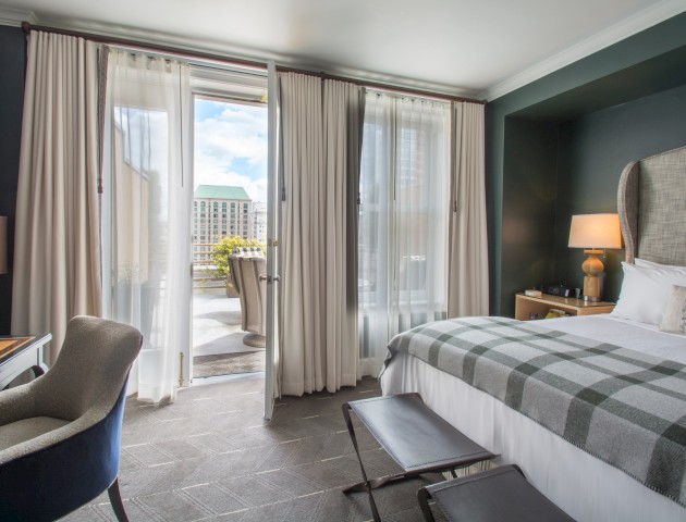 A modern hotel room with a large bed, balcony door, seating area, and city view through sheer curtains.