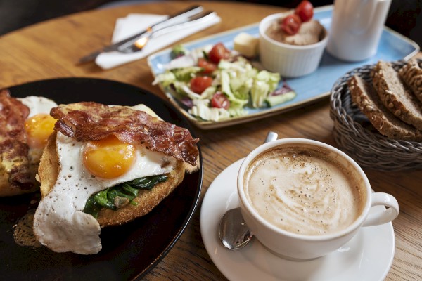 A breakfast with eggs Benedict, bacon, salad, bread, and a frothy coffee on a wooden table.