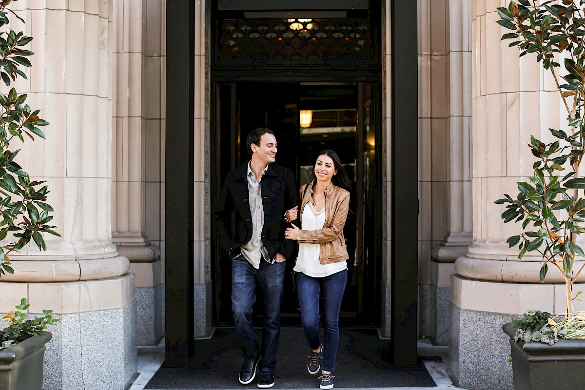 A smiling couple walks arm-in-arm out of a building with stone columns and greenery on either side.