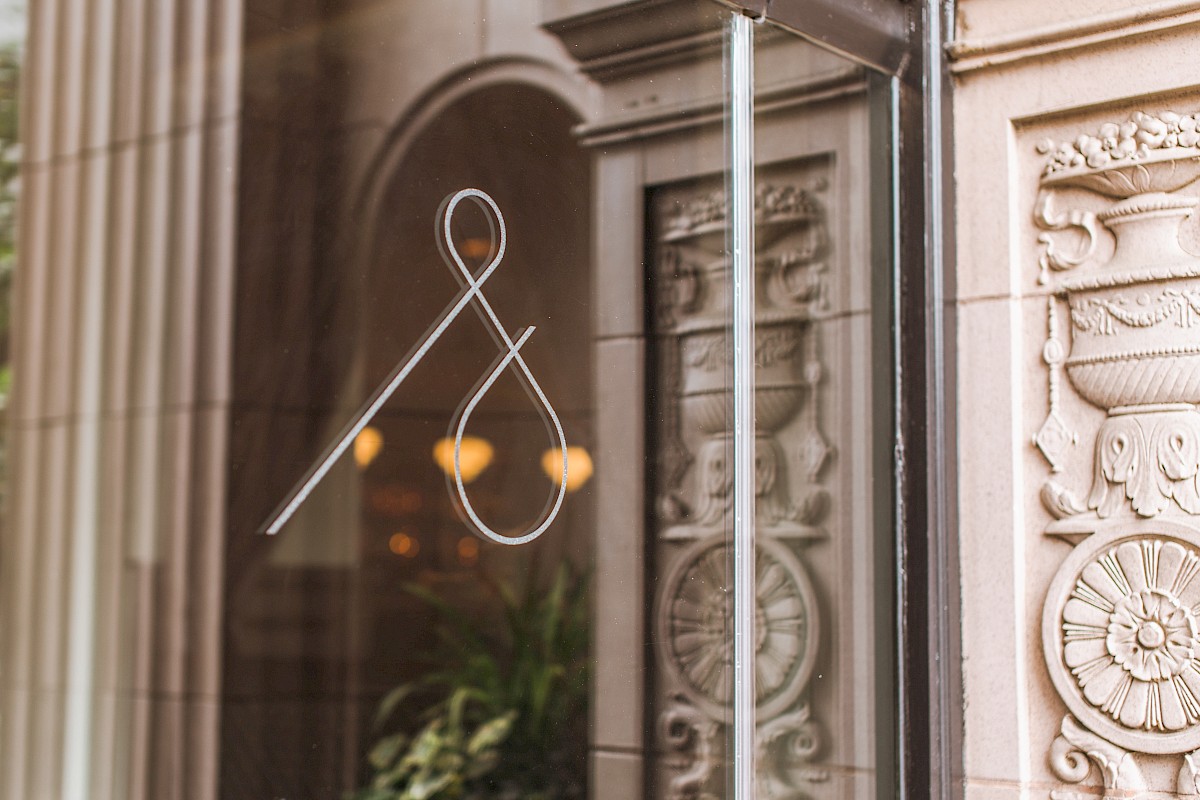 The image shows a glass door with an elegant ampersand (&) symbol, and ornate stone architectural details visible through the glass.