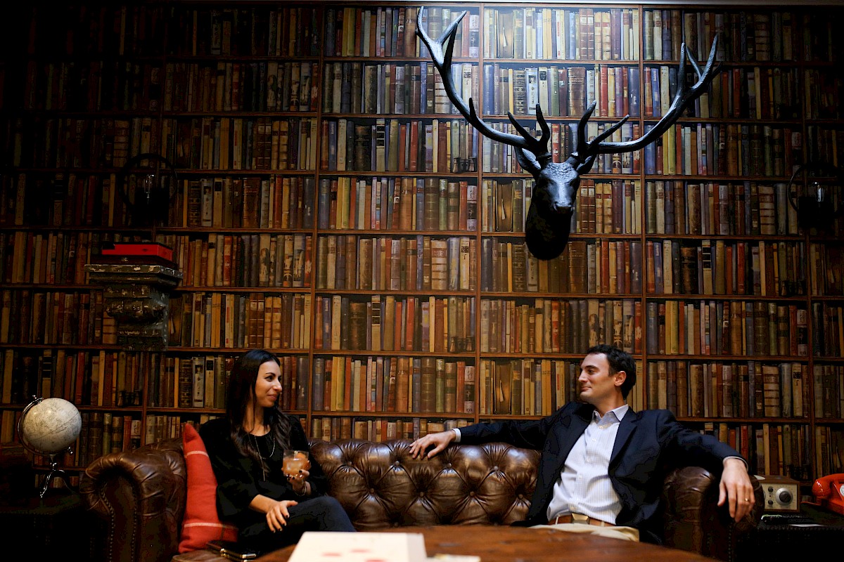 Two people are sitting on a brown leather sofa in front of a large bookshelf wall with a mounted deer head.