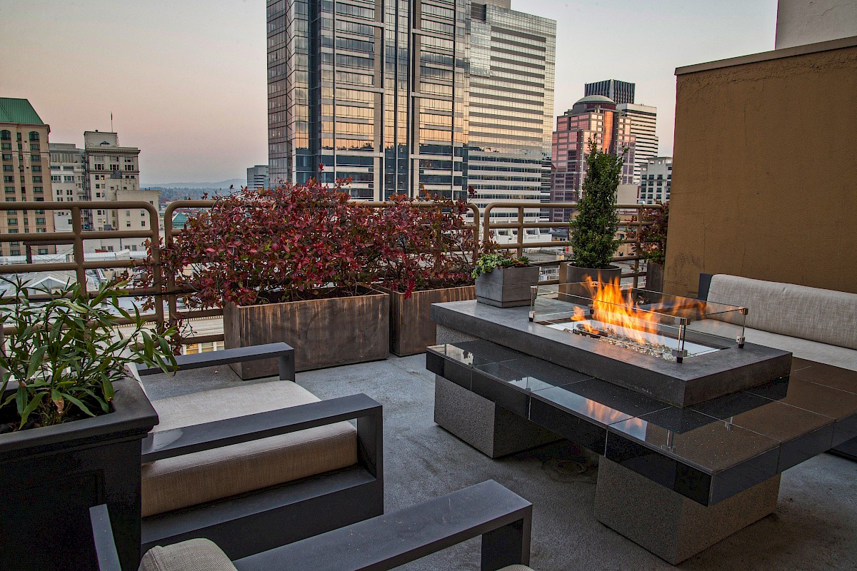 A rooftop patio in a city with modern buildings, outdoor furniture, potted plants, and a fire feature at sunset.