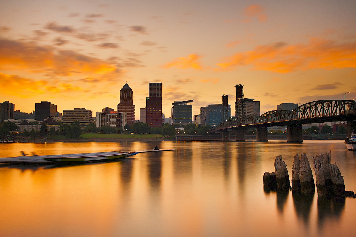 A city skyline at sunset with a river, boat, bridge, and remnants of old wooden posts in the water.