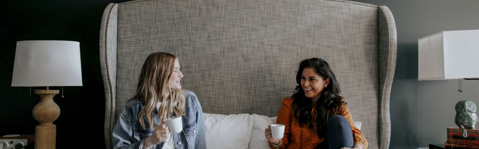 Two women are sitting on a bed, chatting and laughing while holding cups; there are desserts on a tray in front of them.