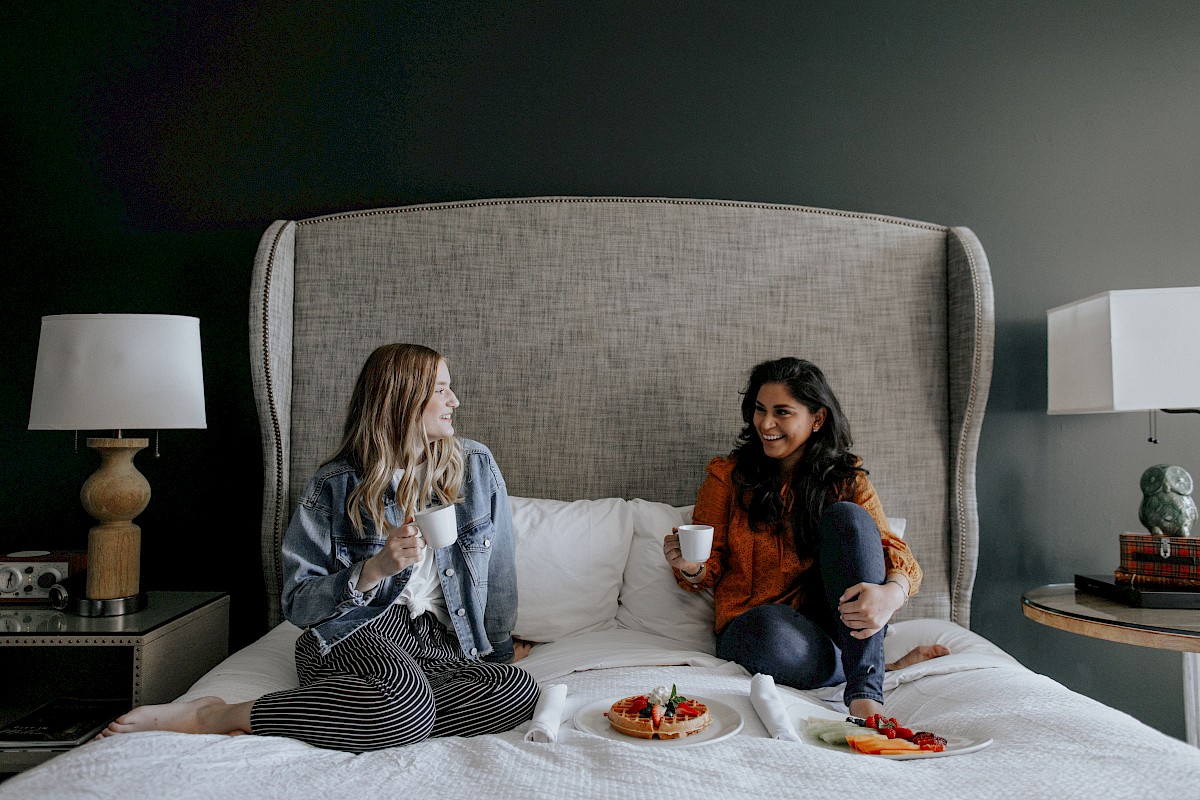 Two women are sitting on a bed, enjoying tea and snacks, with a cake and fruit platter in a cozy, stylish bedroom with dark walls.