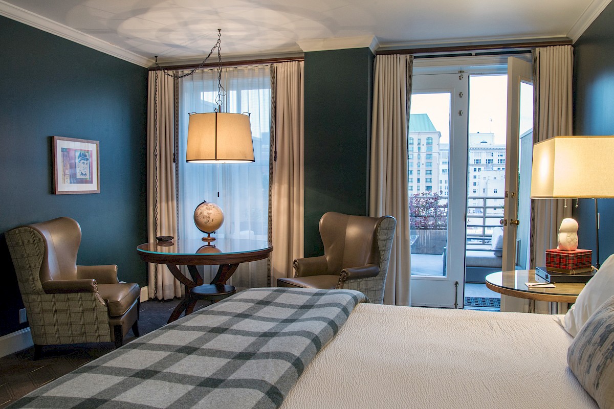 A cozy hotel room with a bed, armchairs, a round table, and a balcony view of city buildings.