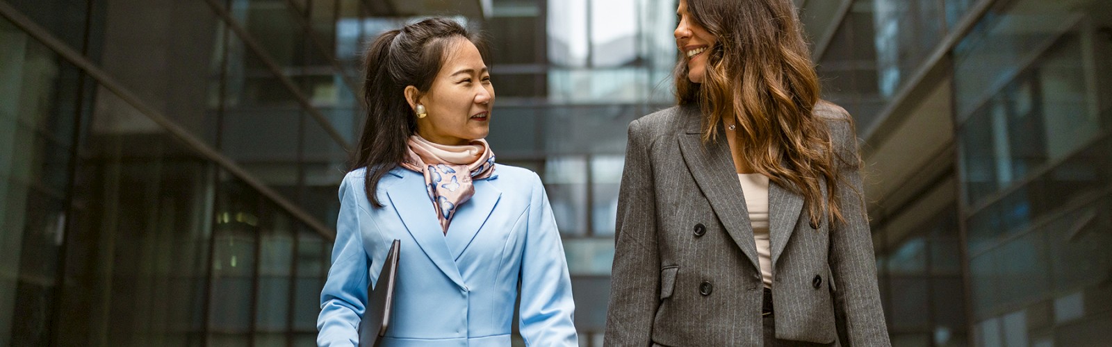 Two women in business attire walk and chat in a modern office building atrium, smiling and holding files or bags.