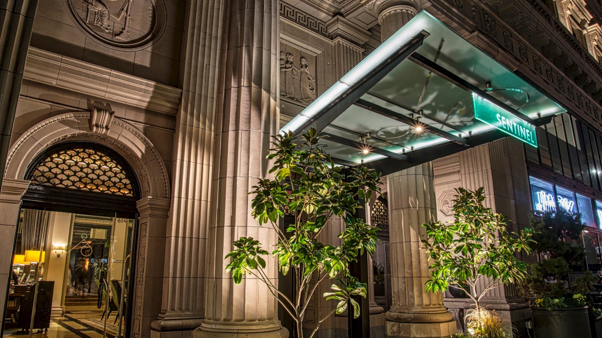 The image shows the entrance of a building with classical architecture, columns, and greenery, featuring neon signs reading 