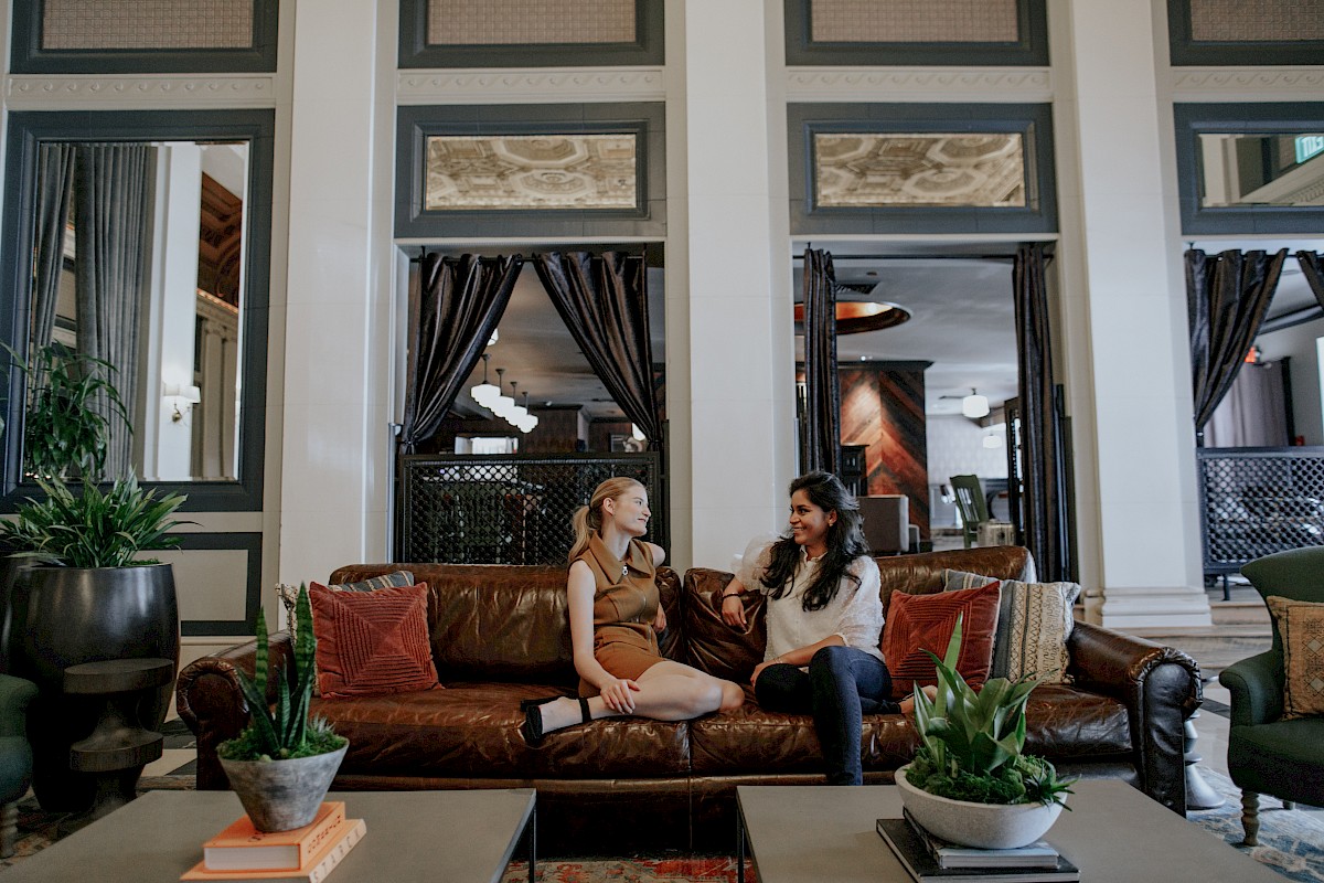 Two women are sitting and chatting on a leather sofa in an elegant, modern hotel lobby filled with plants and stylish decor.