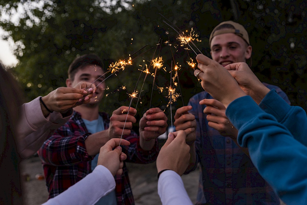 A group of friends is celebrating outdoors with sparklers at dusk, smiling and holding sparklers together in a joyful moment.