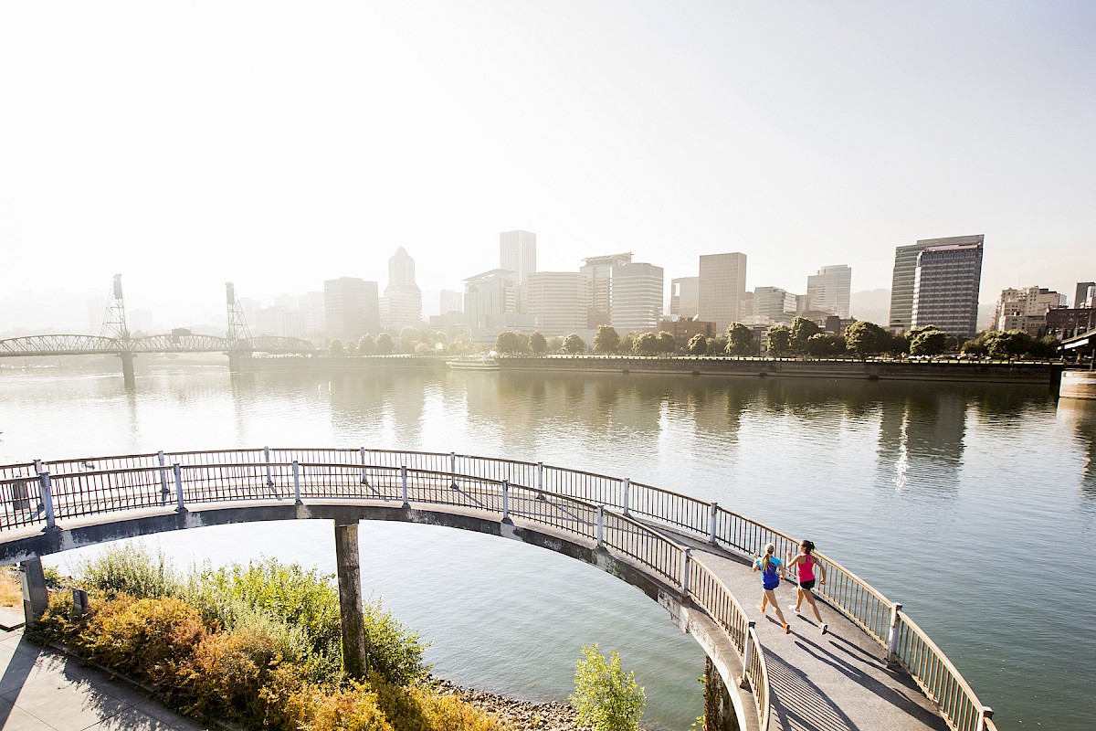 Two women are jogging on a curved bridge by a river with a city skyline in the background.
