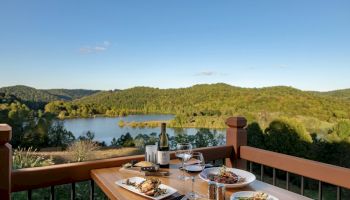 A table set for two with wine and plates of food, overlooking a scenic lake surrounded by hills and greenery under a clear blue sky.