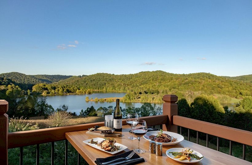 A table set for two with wine and plates of food, overlooking a scenic lake surrounded by hills and greenery under a clear blue sky.