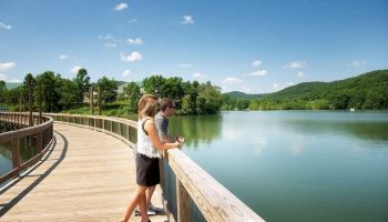 Two people stand on a wooden boardwalk overlooking a serene lake surrounded by lush, green hills under a clear blue sky with scattered clouds.