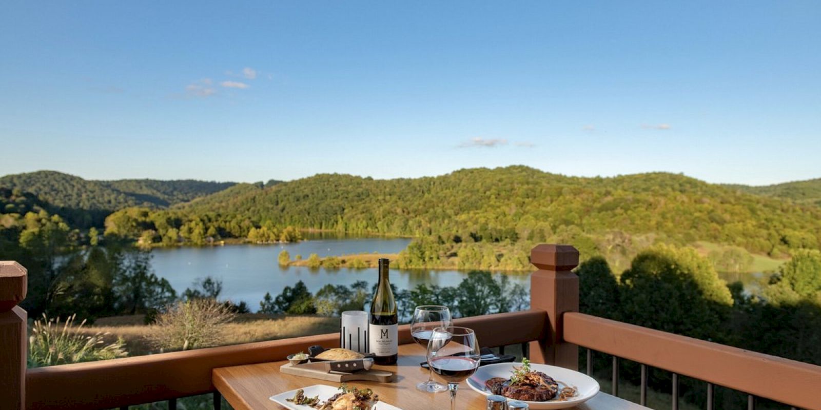 An outdoor dining setup with wine and dishes on a wooden table overlooking a scenic lake and forested hills under a clear blue sky.