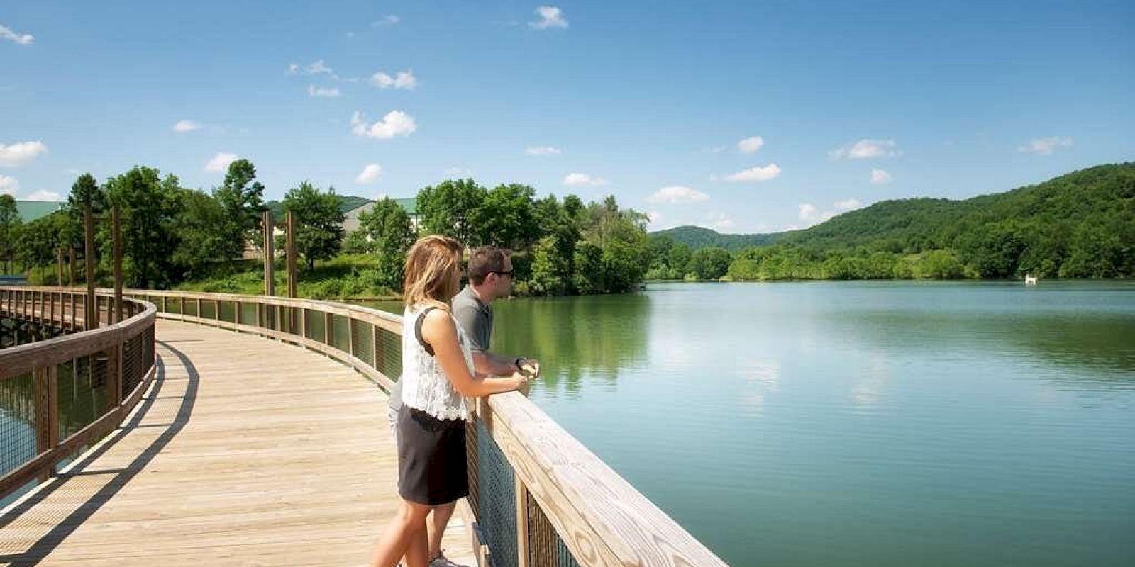 Two people stand on a wooden boardwalk overlooking a serene lake surrounded by lush green hills under a clear, blue sky.