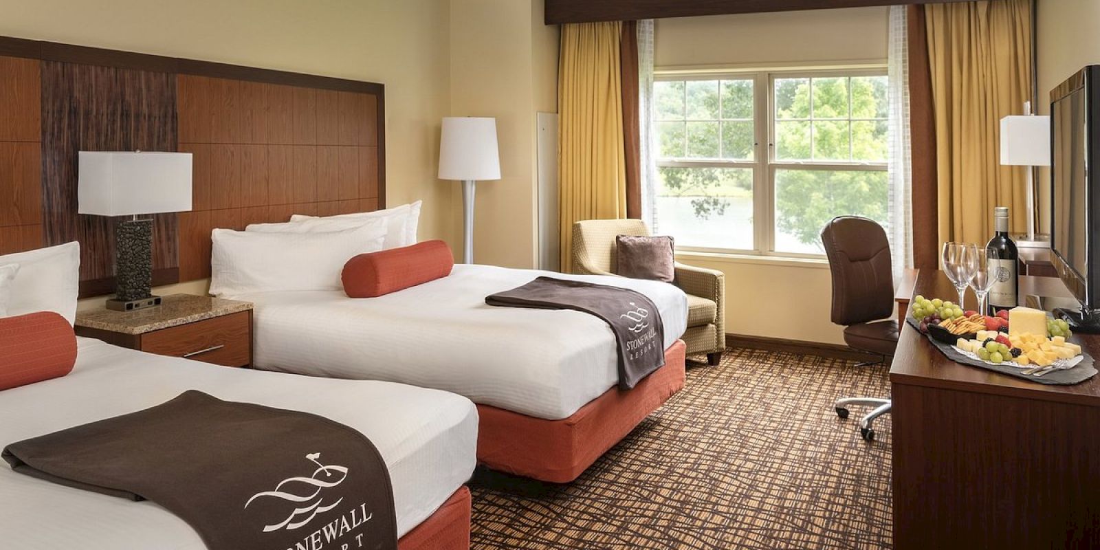 The image shows a hotel room with two double beds, a window with curtains, a TV, a chair, and a table with wine and fruit.