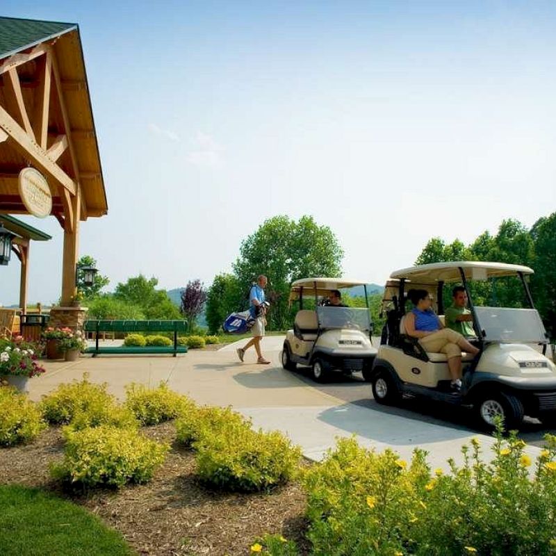 Two golf carts are parked near a building while a person walks carrying golf equipment outside. Lush greenery surrounds the area.