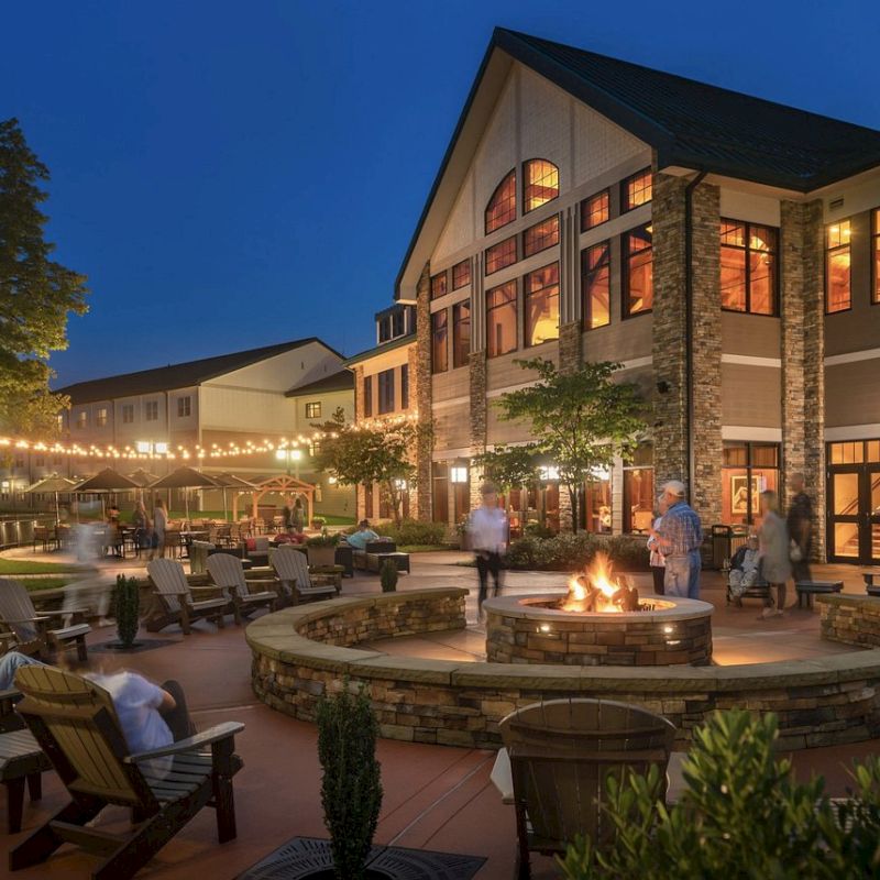 An outdoor gathering space at dusk features a fire pit, cozy seating, string lights, and a large building in the background, with people relaxing.