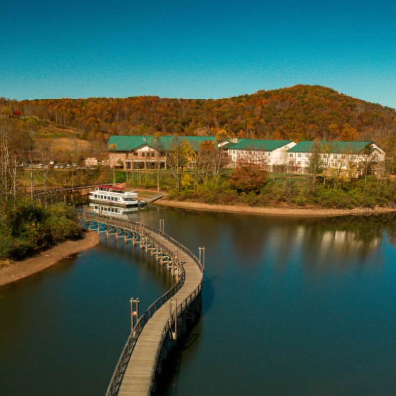An aerial view of a serene lake with a curved walkway leading to a structure surrounded by trees, hills in the background, and clear skies.