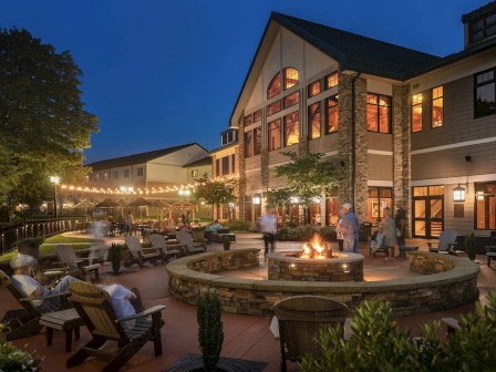 Evening scene of a lit-up outdoor patio with a fire pit, surrounded by chairs, people relaxing, and a large building in the background.