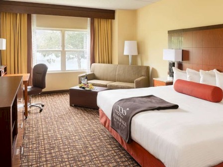 A hotel room with a bed, sofa, desk, chair, TV, lamps, and a window with curtains. The bed has white sheets and a blanket with a logo.