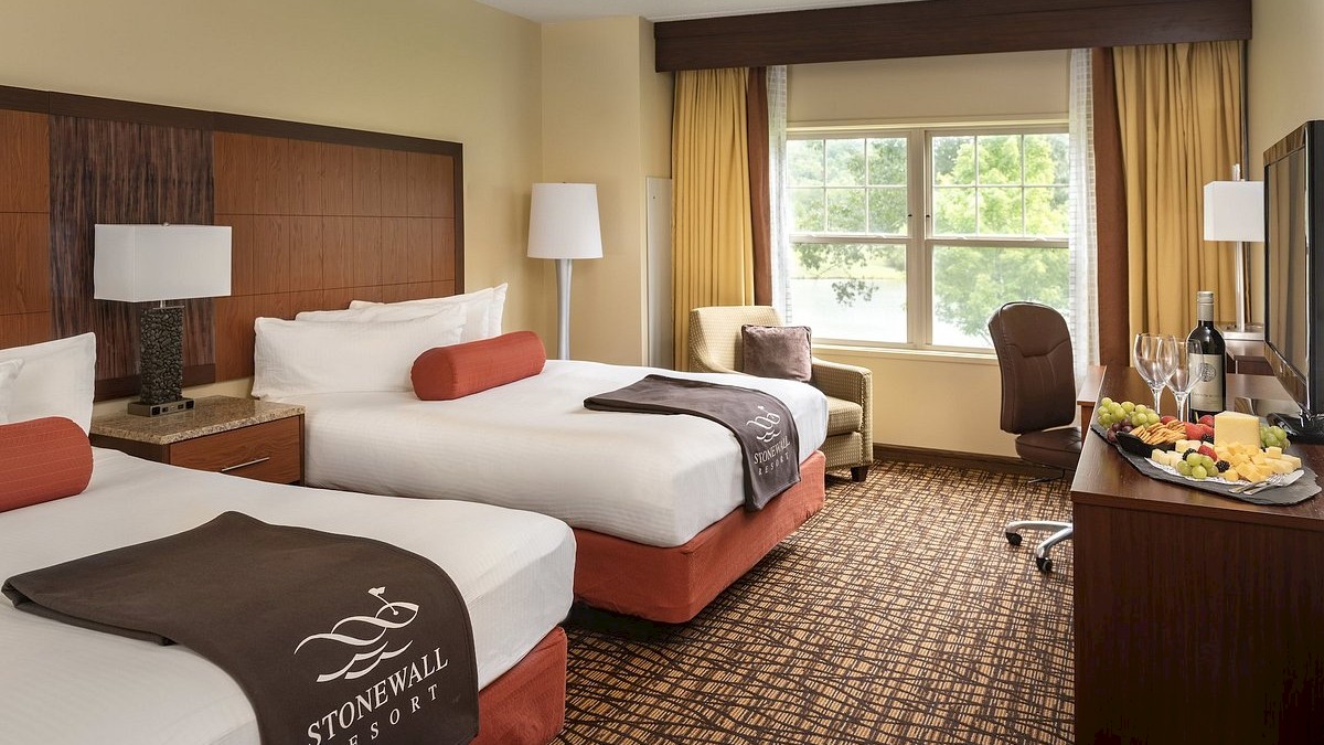Two double beds with Stonewall Resort logos, a chair, lamps, a window, and a TV. A wine bottle and fruit plate are on the dresser.