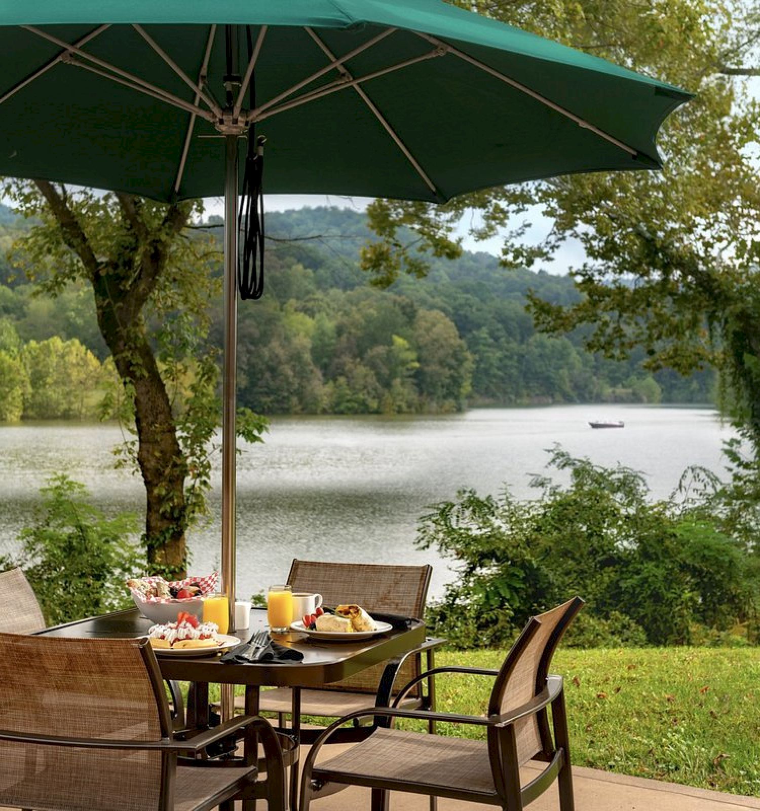 A scenic outdoor dining area with a green umbrella, table, chairs, and plates of food overlooking a peaceful lake and lush, green trees ending the sentence.