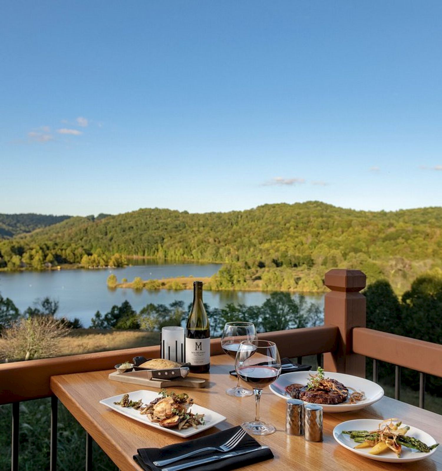 A scenic outdoor dining setup with a table, wine, and plates of food overlooking a lake and forested hills under a clear sky.
