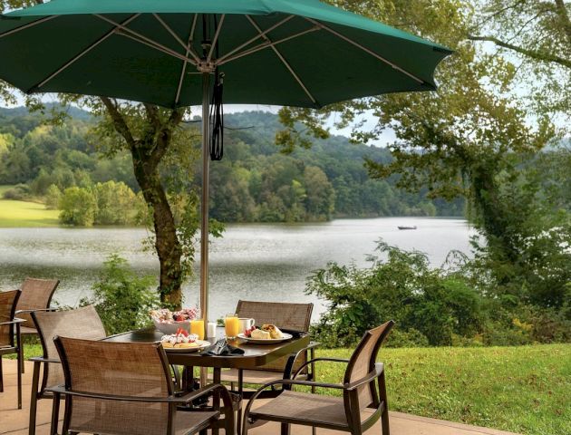 An outdoor dining setup with a green umbrella, chairs, and a table set with food, overlooking a scenic lake surrounded by trees and hills.