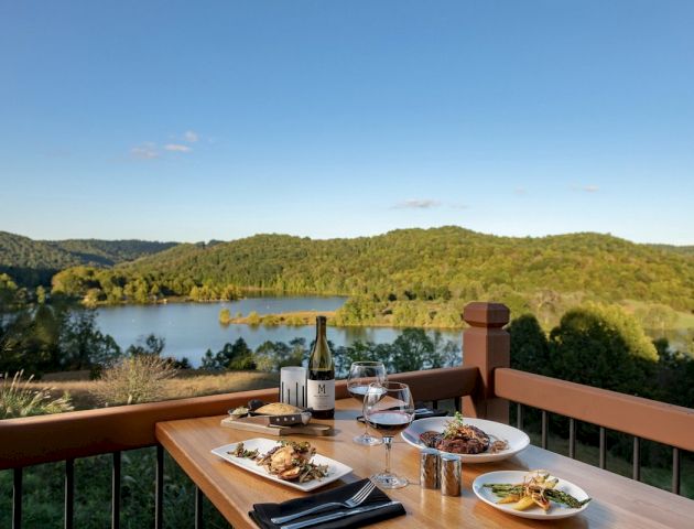 A wooden table with meals, a wine bottle, and glasses overlooks a scenic landscape with a lake and green hills on a clear day.