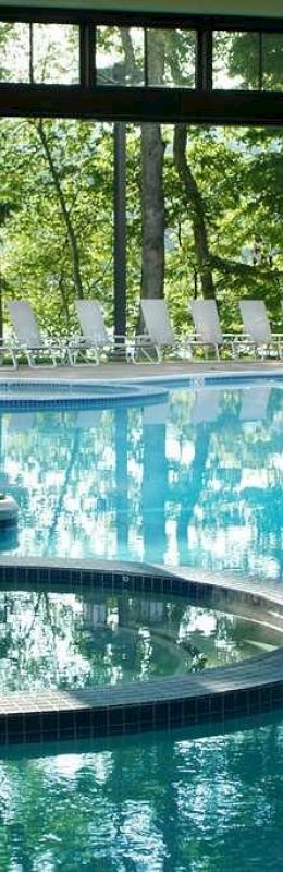 An indoor pool area with windows showing an outdoor pool and trees, featuring clear blue water and lounge chairs outside.