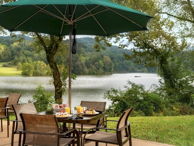 An outdoor dining table with food and drinks, under a green umbrella, with a scenic view of a lake surrounded by trees and hills.