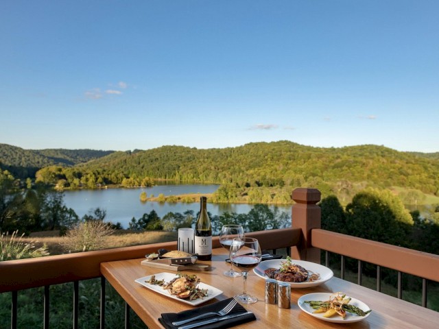 A table set with plates of food, wine, and glasses on a balcony overlooking a scenic landscape with a lake and green hills under a clear sky.
