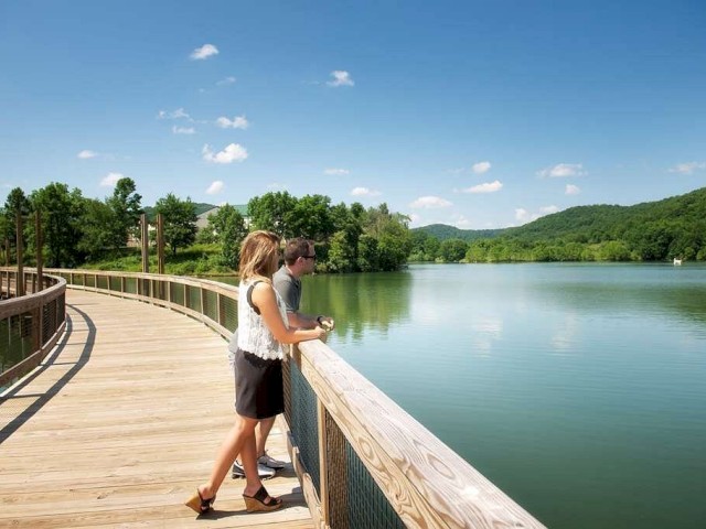Two people stand on a wooden boardwalk overlooking a calm lake surrounded by greenery and hills under a clear, blue sky.