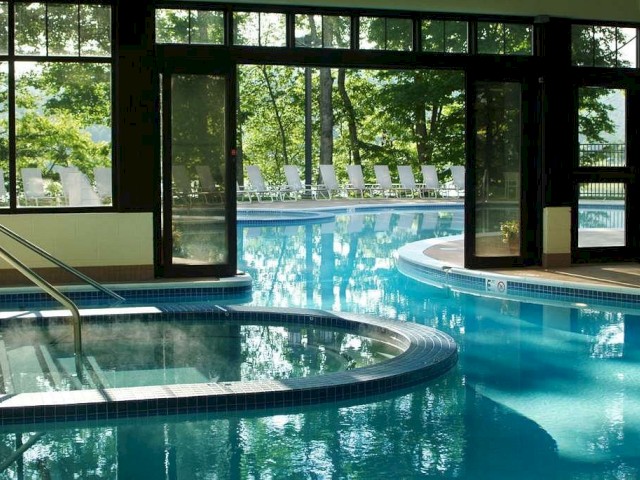 This image shows an indoor-outdoor pool area with large windows, a relaxing hot tub, and loungers under the trees, with sunlight streaming in.