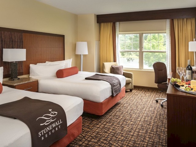 A hotel room with two double beds, a window, a small sitting area, and a desk with a fruit platter and wine, featuring Stonewall Resort amenities.