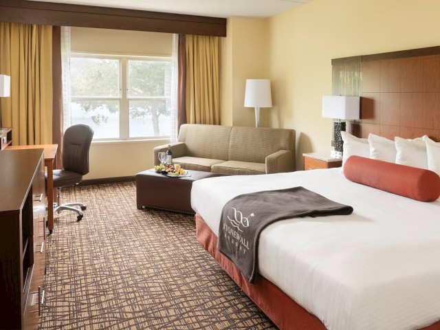 A hotel room with a large bed, couch, desk, chair, TV, and large window. The room is well-lit and decorated in neutral tones, ending the sentence.