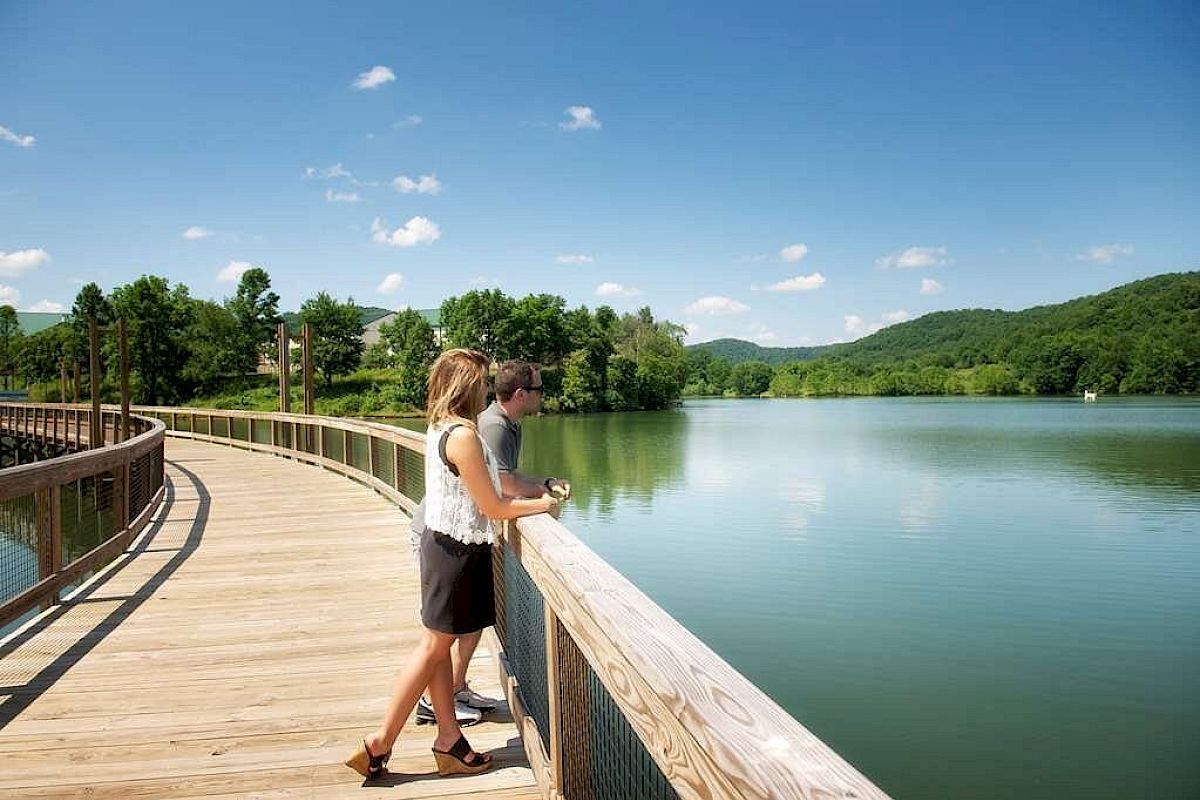 A man and a woman are standing on a wooden boardwalk, overlooking a peaceful lake surrounded by lush green hills under a clear blue sky, ending the sentence.