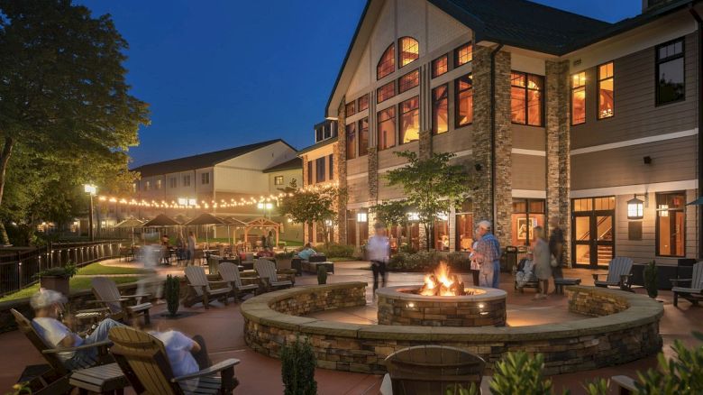 An outdoor area with a fire pit, string lights, and seated people at a resort or hotel in the evening; the building is lit with warm lights.